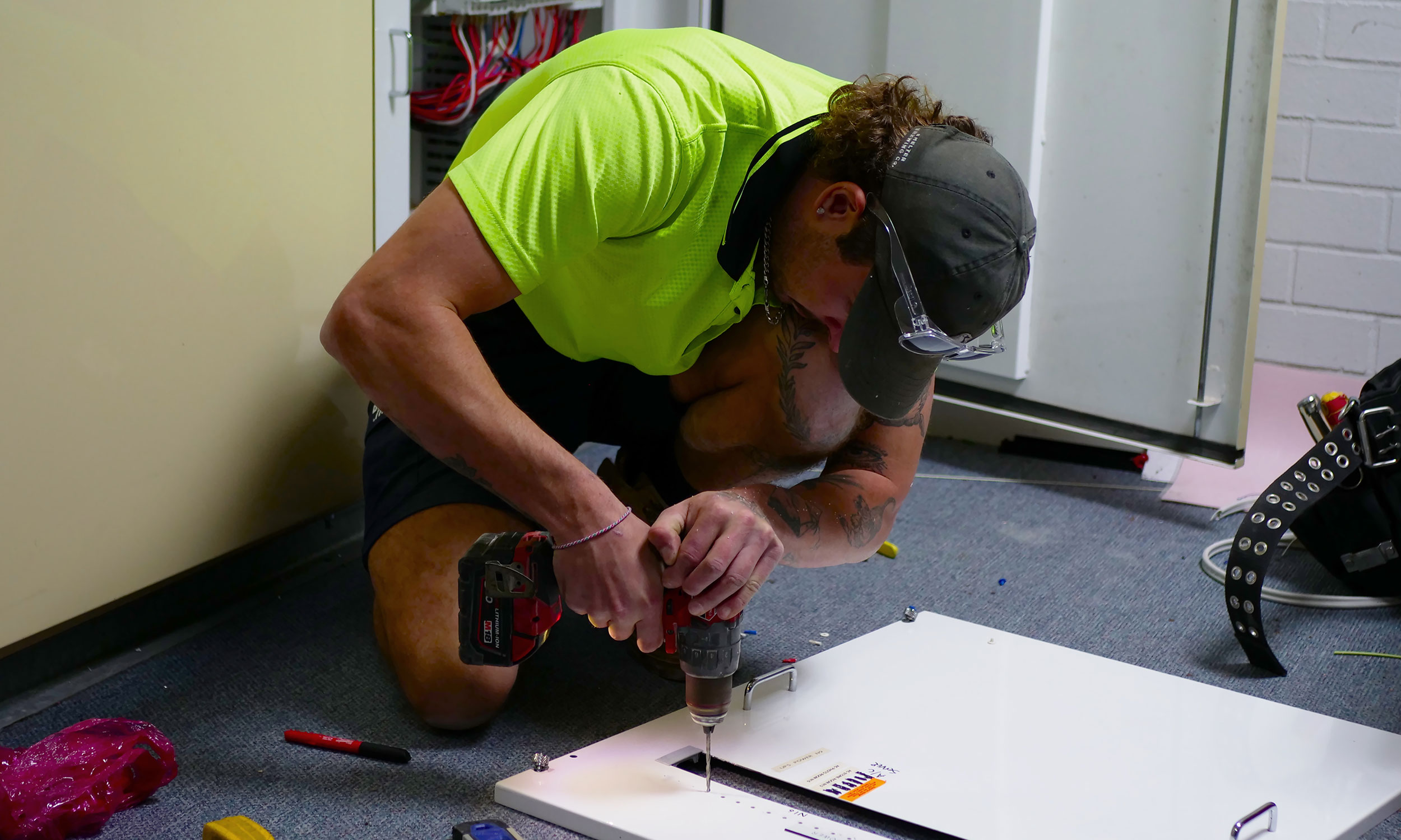 A man in a green shirt and a hat repairs a refrigerator, showcasing his skills as an electrician in Canning Vale