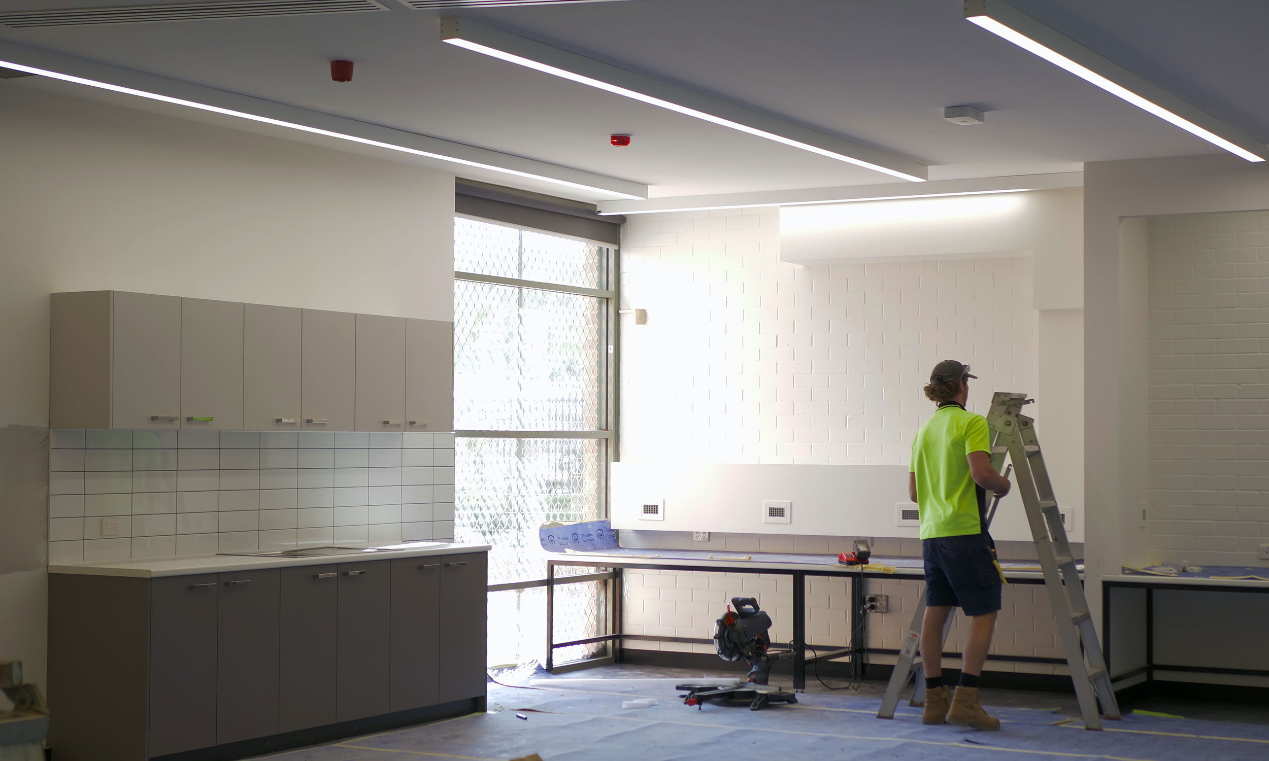 A commercial electrician in Perth wears a green shirt and cap stands in a kitchen holding a ladder, ready to begin electrical work